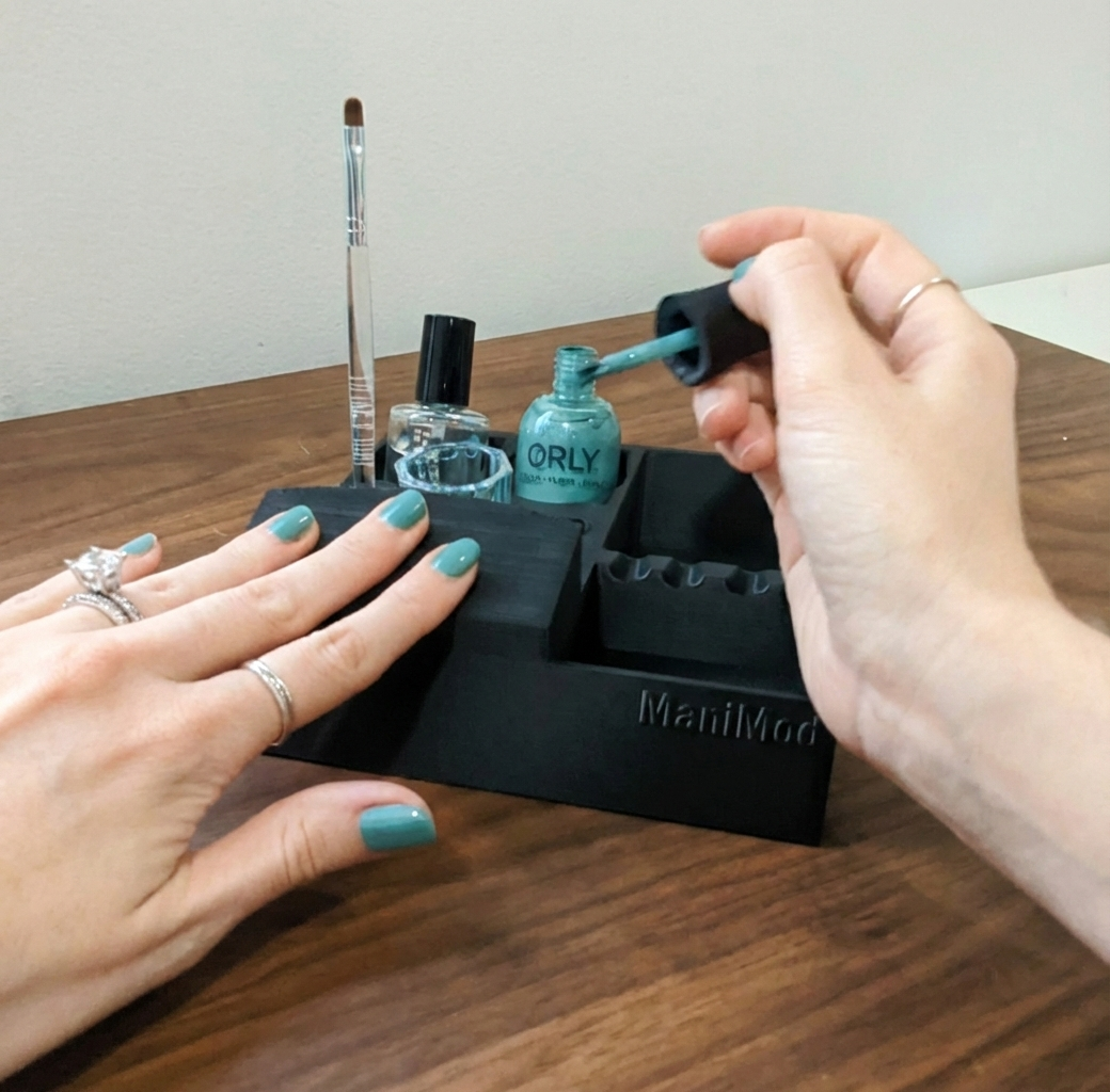 "Manimod XL black nail polish working tray holding a teal Orly bottle, a glass dappen dish, and a manicure brush, featuring a built-in hand rest for at-home nail care."